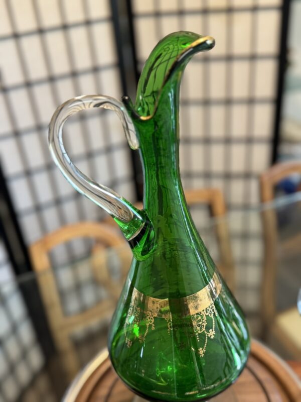 A green glass decanter with a gold decorative band and a clear handle sits on a round wooden tray. The decanter is photographed indoors, in front of wooden chairs and a black-and-white folding screen.