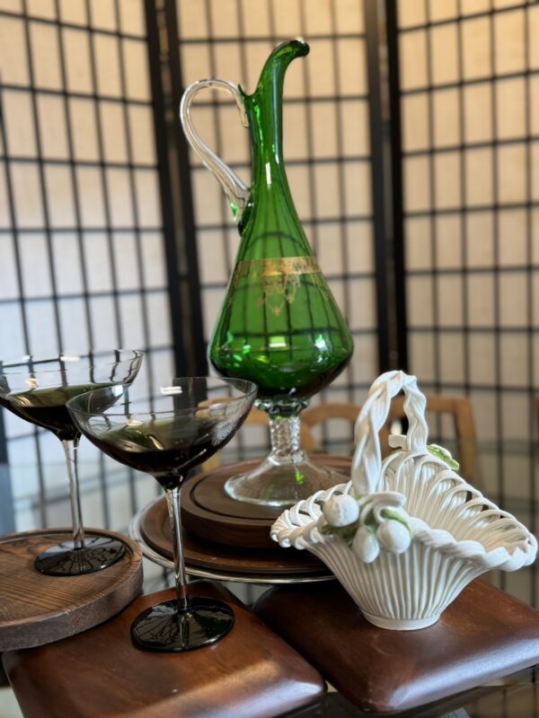 A tall, green glass decanter with gold accents stands on a table beside two filled cocktail glasses on round wooden coasters. A white ceramic basket with sculpted flowers sits in the foreground. A folding screen serves as the background.