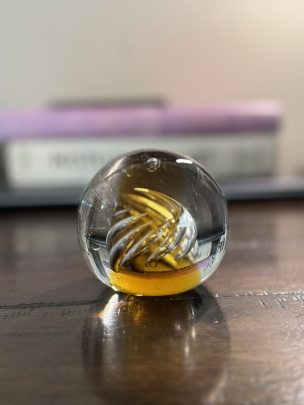 A clear glass paperweight with a yellow and white spiral design inside sits on a wooden surface. In the blurred background, stacked books with a purple cover on top are partially visible. Warm lighting highlights the smooth glass.
