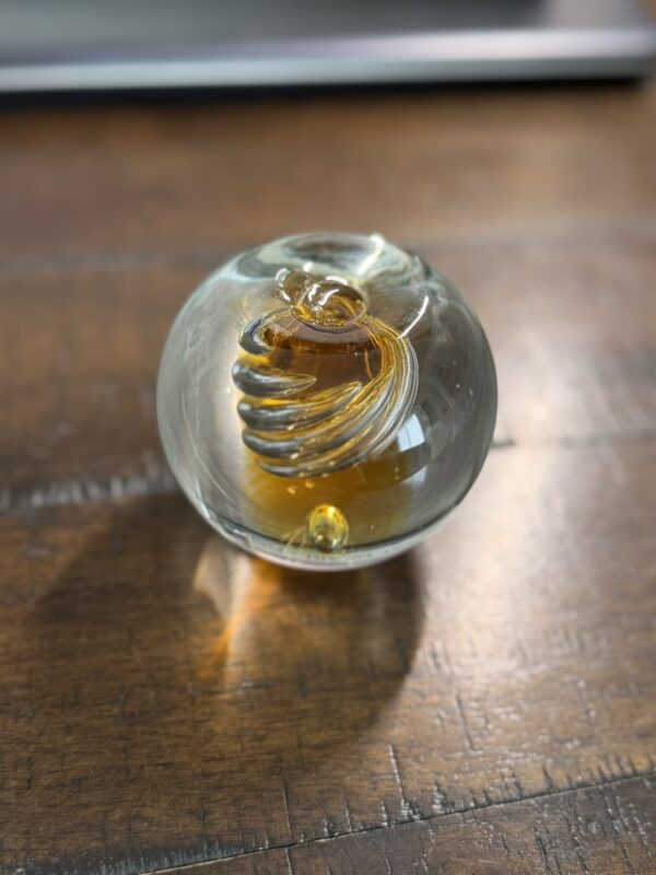 A clear glass sphere with an amber spiral design inside rests on a wooden surface. Sunlight highlights the glass, casting soft reflections and shadows. The background is slightly out of focus, emphasizing the intricate details of the sphere.