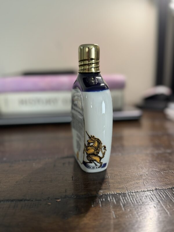 A small bottle with a gold cap stands on a wooden table. The white bottle features a blue heraldic unicorn design. In the blurred background are stacked books and some indistinct objects, creating a cozy, softly lit ambiance.