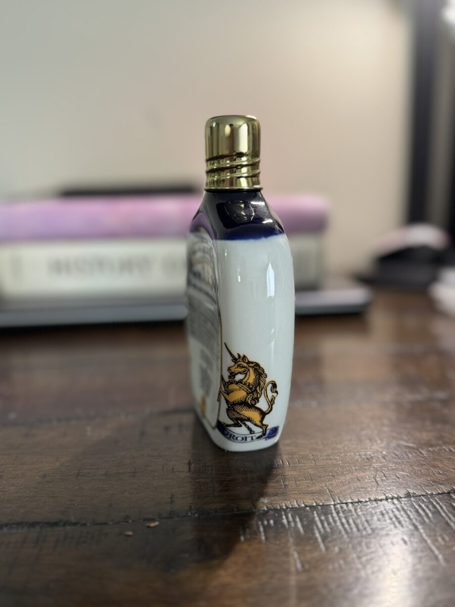 A small bottle with a gold cap stands on a wooden table. The white bottle features a blue heraldic unicorn design. In the blurred background are stacked books and some indistinct objects, creating a cozy, softly lit ambiance.