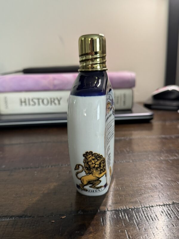A decorative bottle with a gold lion emblem and gold cap stands on a wooden table. Behind it, a closed laptop, two stacked books—one titled HISTORY—and a computer mouse are visible. The background is softly lit.