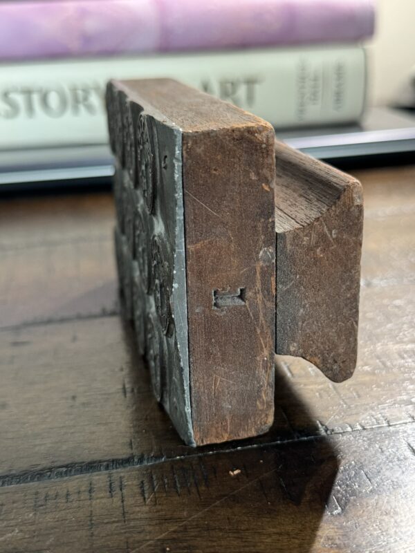 A close-up side view of the Antique Clock Printing Block with detailed metalwork on one side, resting on wood. In the blurred background, books are stacked horizontally, showing STORY and ART on their spines.