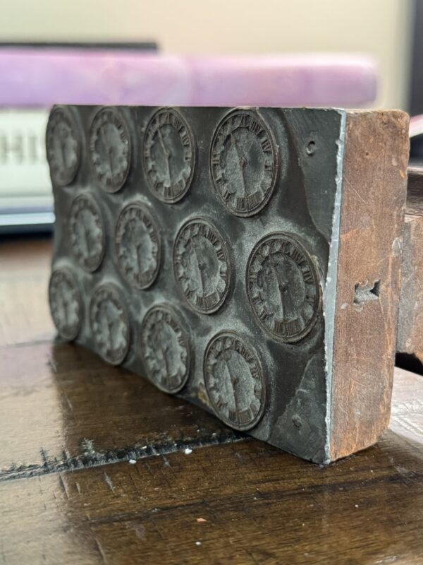 A close-up of the Antique Clock Printing Block, a rectangular metal and wood mold with twelve circular profile impressions framed by Roman numerals, resting on a wooden surface with blurred background objects.