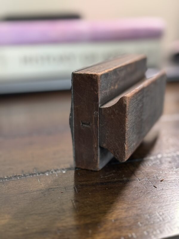 A close-up of the Antique Clock Printing Block, a vintage rectangular wood and metal piece with a grooved center, resting on a wooden surface with blurred stacks of books in the background.