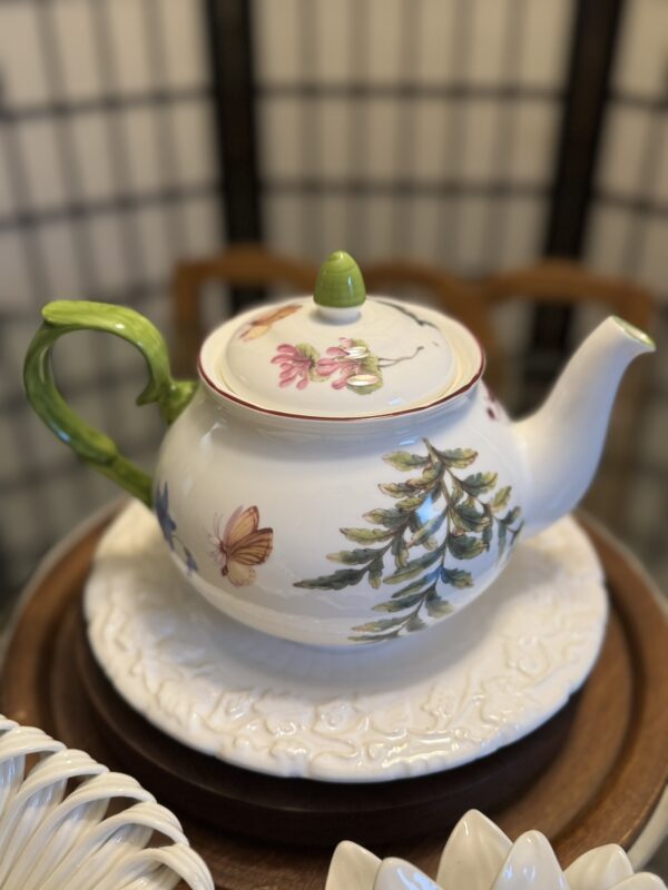 A white ceramic teapot with a green handle and knob, decorated with painted flowers, leaves, and a butterfly, sits on an ornate white plate. The background is softly blurred, showing wooden furniture and a dark grid-patterned screen.