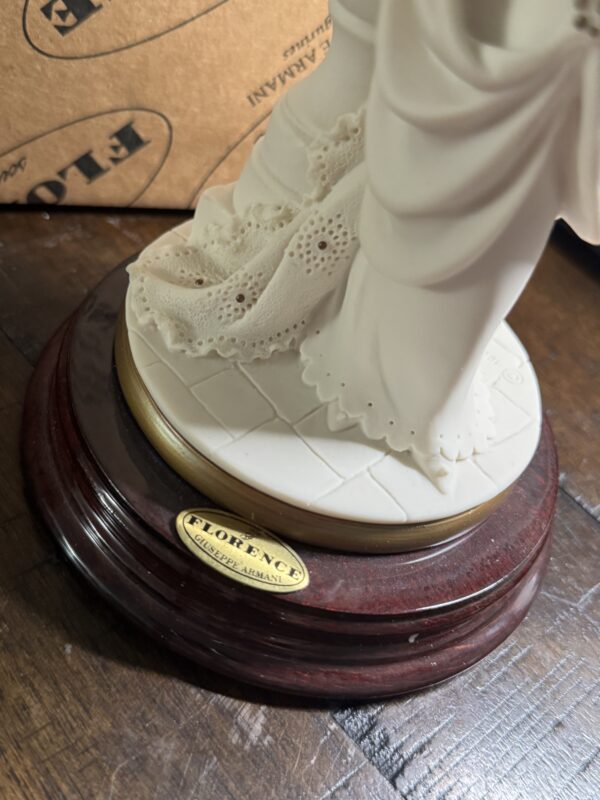 A close-up of the Armani Bride Figurine shows detailed lace on the dress and a round wooden base with a gold FLORENCE GIUSEPPE ARMANI plaque. The figurine is on white tiles, with its cardboard box visible in the background.