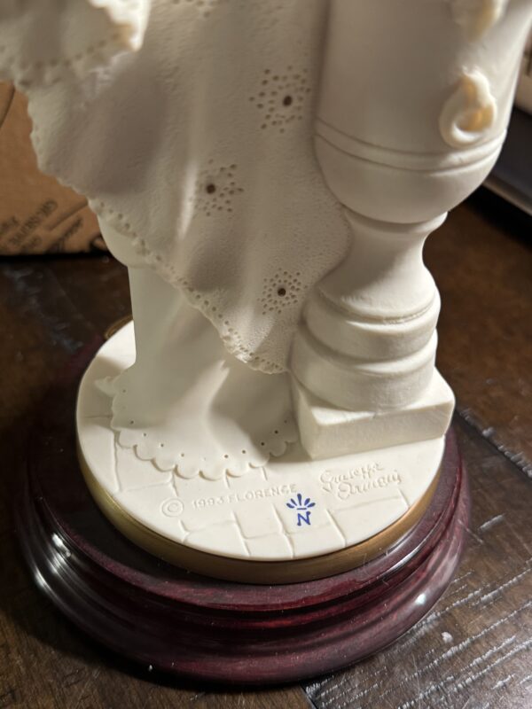 Close-up of the Armani Bride Figurine's white porcelain base on a dark wooden surface, featuring © 1995 FLORENCE, Giuseppe Armani marks, a blue N mark, and lace-like details along the robe edge.