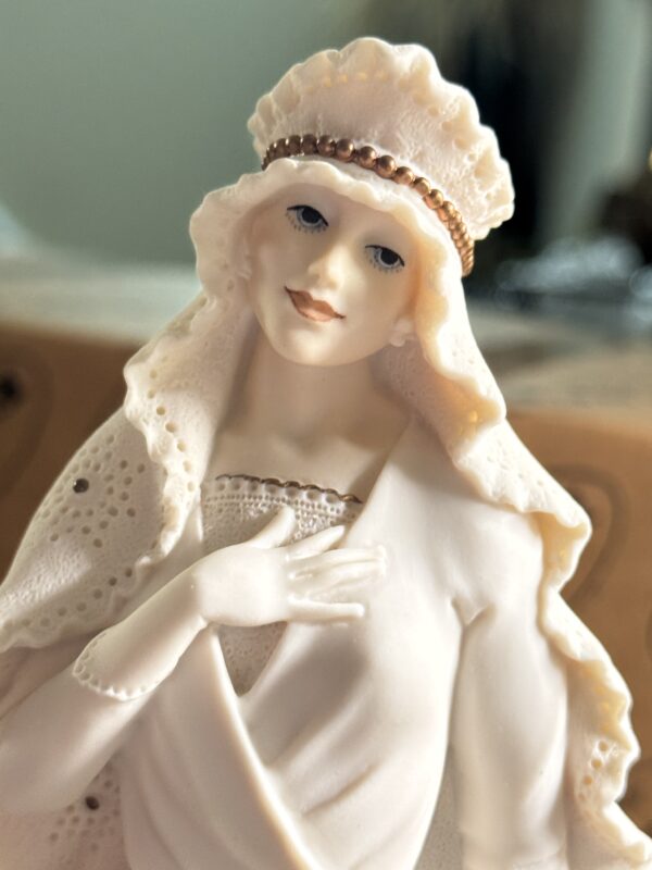 A close-up of the Armani Bride Figurine reveals a porcelain woman with pale skin, blue eyes, and light lips. She wears a textured white headdress with a gold band, a matching gown, and holds her hand gracefully to her chest.