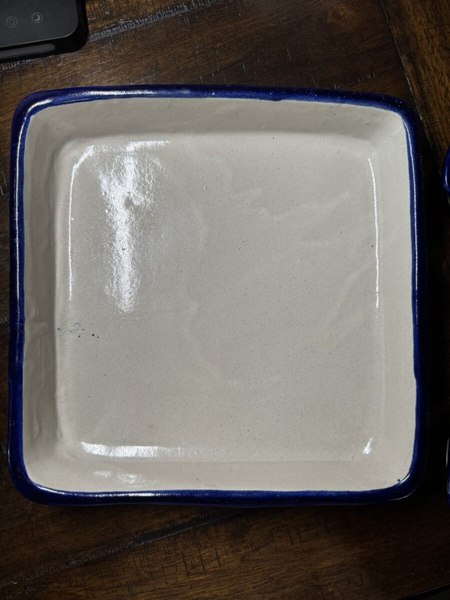 A square ceramic baking dish with a shiny beige interior and a dark blue rim sits on a wooden surface. The dish has rounded corners and a smooth, glossy finish, reflecting light on one side. A small part of another object is visible in the top left corner.
