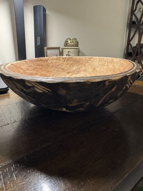 A large, shallow wooden bowl with a dark exterior adorned with carved leaf patterns and a lighter, textured interior sits on a wooden table. In the background are decorative items, including a gold container and framed pictures.