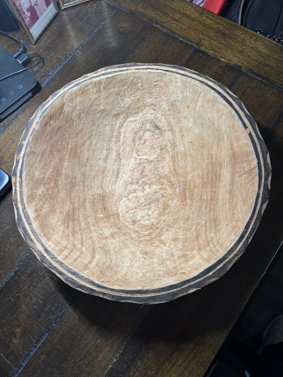 A round, shallow wooden bowl with a textured, natural grain and a rough, darkened rim sits on a wooden table. The surface of the bowl is unfinished, and parts of a smartphone and framed photo are visible at the edges of the image.