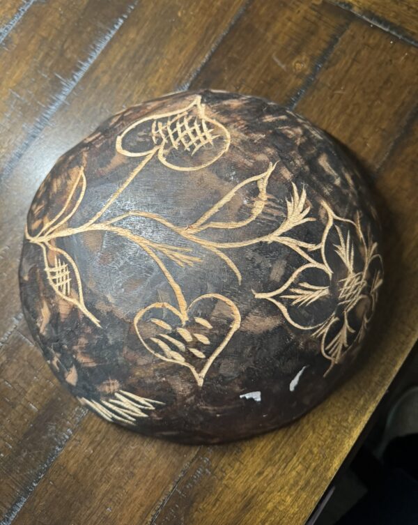 A round, dark brown, coconut shell bowl rests on a wooden surface. The bowl features hand-carved floral and heart designs etched in light tan lines, creating a contrast with the dark background. The carvings give the bowl a rustic, handmade appearance.
