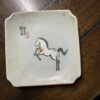 A square ceramic plate with crackled glazing features a painted black and white horse rearing on its hind legs. There are two red stamps and black Chinese calligraphy in the top left corner. The plate rests on a brown wooden surface.