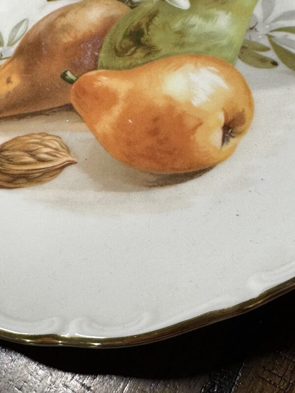 A close-up of a decorative plate with a scalloped, gold-trimmed edge, featuring a painted design of a yellow-orange pear, a brown nut, parts of two other fruits, and green leaves against a cream background. The plate rests on a wooden surface.
