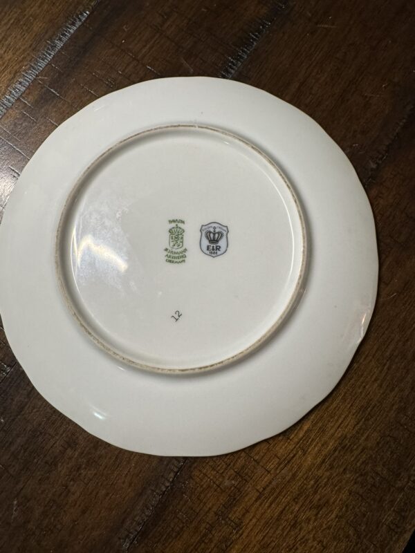 A white ceramic plate is shown upside down on a wooden surface. The plate’s underside has two stamps: one green with a crest and “Rosenthal Selb Bavaria,” and one blue with a shield and crown labeled “EAR.” The plates edge is slightly scalloped.