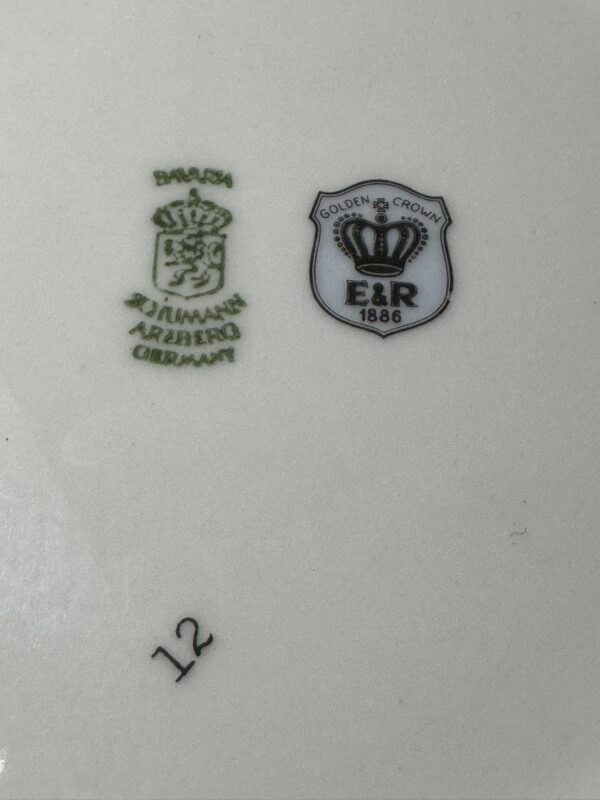 A close-up of a porcelain mark shows two logos: one green with a crown and Bavaria Schumann Arzberg Germany, and one black and white shield with a crown, Golden Crown E&R 1886. The number 12 appears in black below the logos.