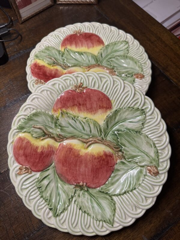 Two decorative ceramic plates with embossed designs of red apples and green leaves, featuring a cream-colored ornate border, are placed overlapping each other on a dark wooden surface. The plates have a glossy, hand-painted appearance.