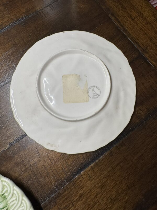 The image shows the back of a white ceramic plate with a wavy edge, placed on a dark wooden table. The plate has a rectangular, worn adhesive label and a faint circular stamp near the center. Some dirt or stains are visible near the plate’s rim.
