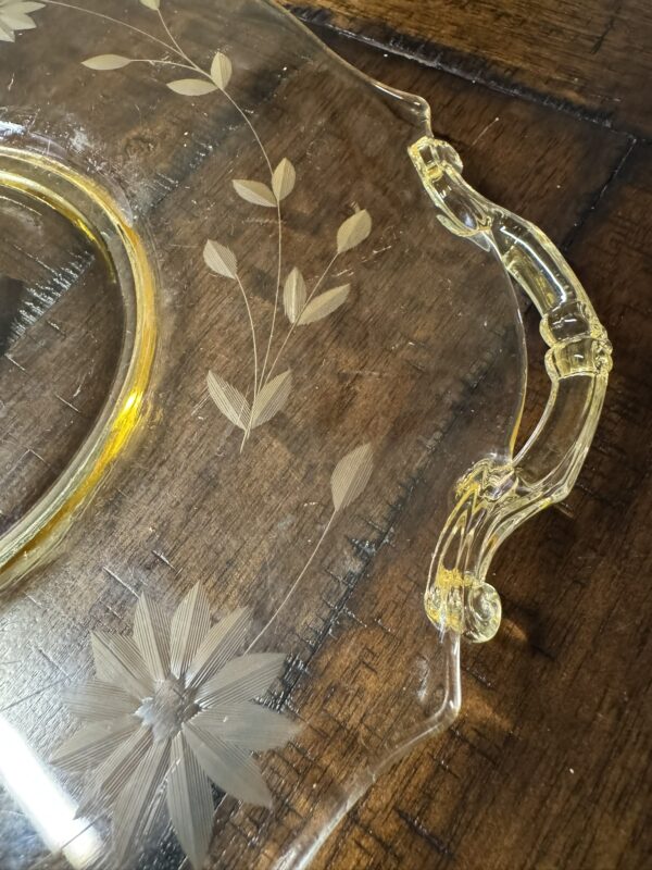 A close-up of a clear glass platter with ornate etched floral and leaf designs. The platter has a decorative handle and sits on a dark wooden surface, showing intricate patterns and a slight yellow tint on the glass edge and handle.
