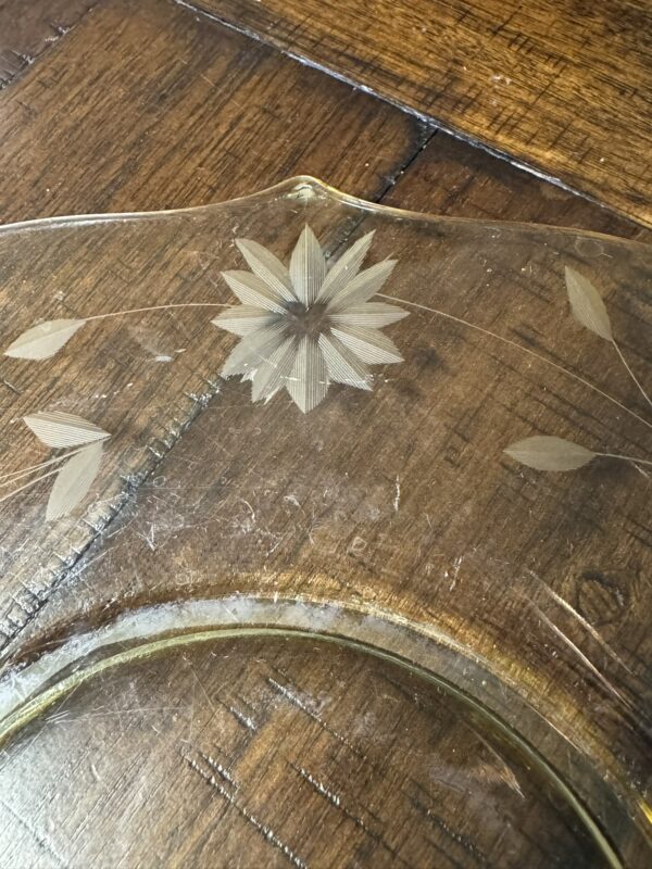 A close-up image of a clear glass plate with an etched floral pattern, featuring a central flower and small leaves. The plate rests on a wooden surface with visible grain and seams, and the glass shows slight wear and scratches.