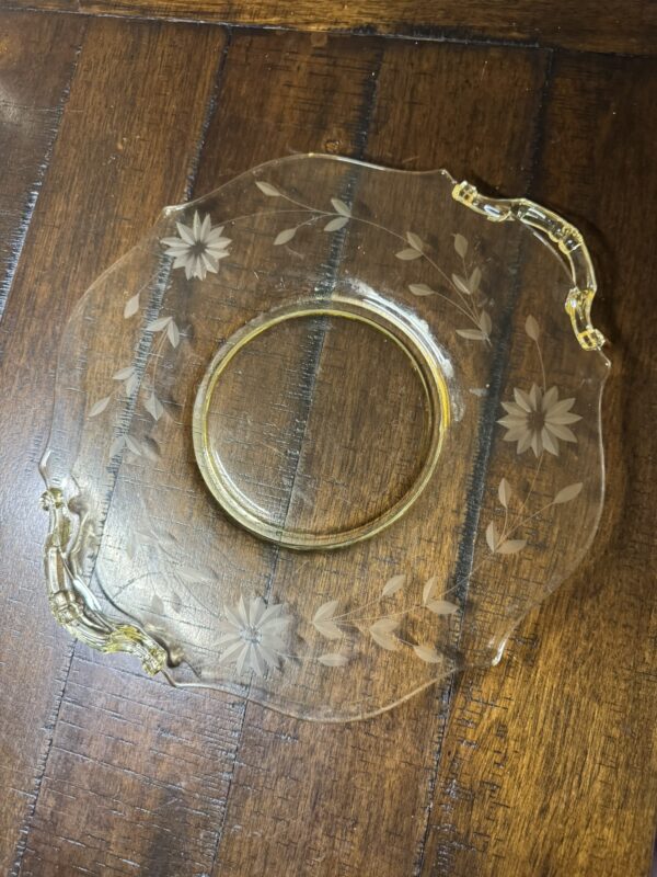 A clear glass plate with two gold-trimmed handles and etched floral designs sits on a dark wooden surface. The plate has a round center with a wide, slightly scalloped rim, decorated with etched flowers and leaves.