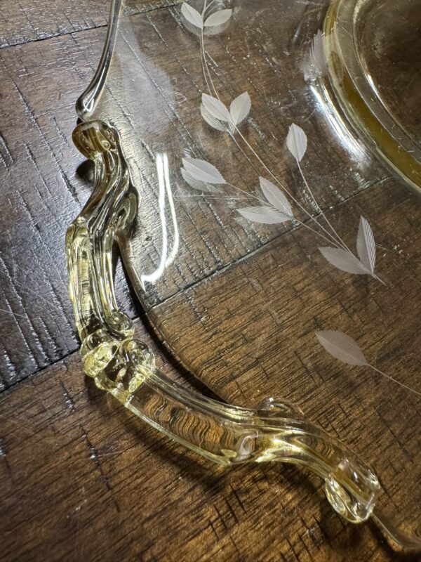 Close-up of a clear glass platter with a curved, yellow-tinted handle resting on a wooden surface. The platter is decorated with etched leafy fern patterns, and the wood grain is visible underneath. Light reflections highlight the glass texture.