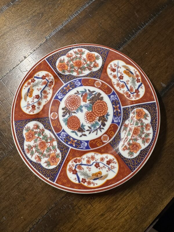 A round decorative plate with intricate floral and bird patterns in red, blue, and gold. The plate has a central flower design, surrounded by alternating sections featuring flowers and birds, all bordered with ornate blue and gold detailing. The plate sits on a wooden table.