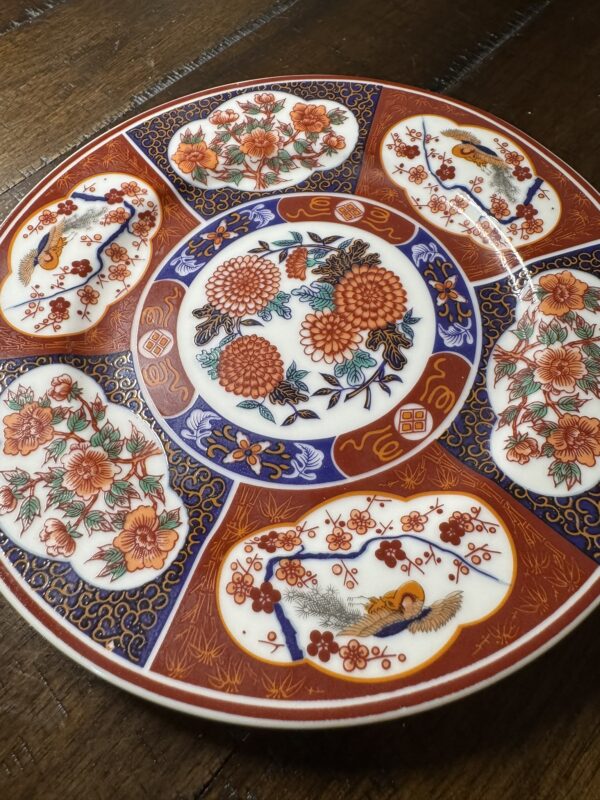 A decorative round plate with intricate red, blue, and gold patterns. The plate features six outer oval sections with floral and bird designs, and a central circular area with detailed orange flowers, all bordered by elaborate geometric and floral motifs.