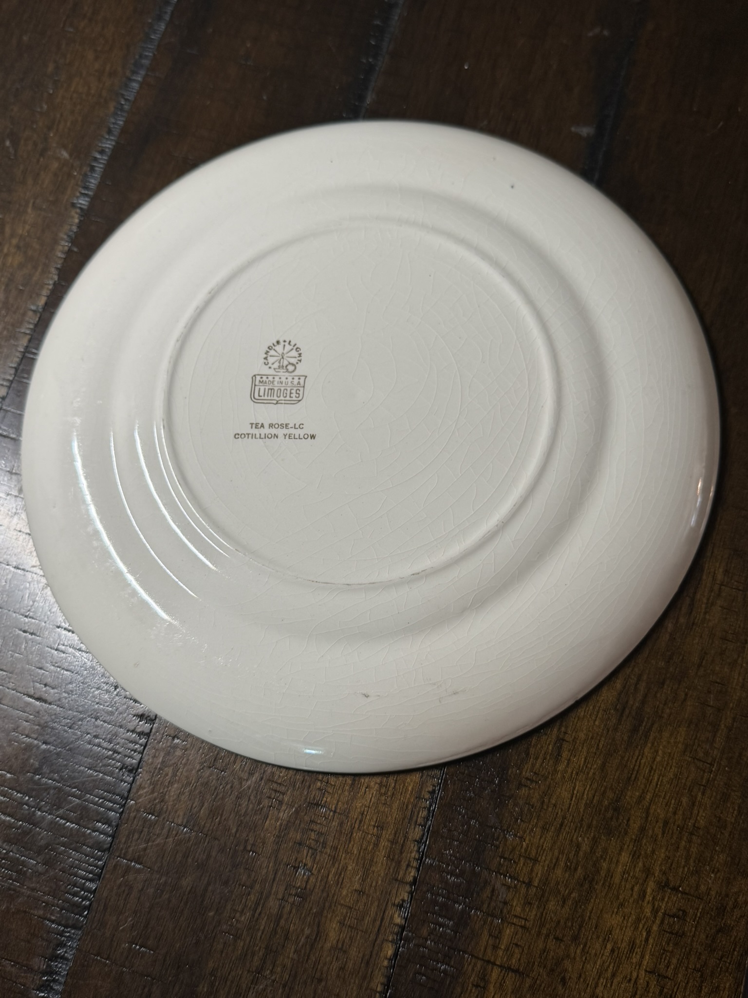 A cream-colored ceramic saucer, seen upside down on a dark wooden surface. The saucer has fine crackle lines and a logo with text at the center, reading: TEA ROSE 4-40, OVENPROOF, YELLOW, USA. Two concentric rings circle the base.