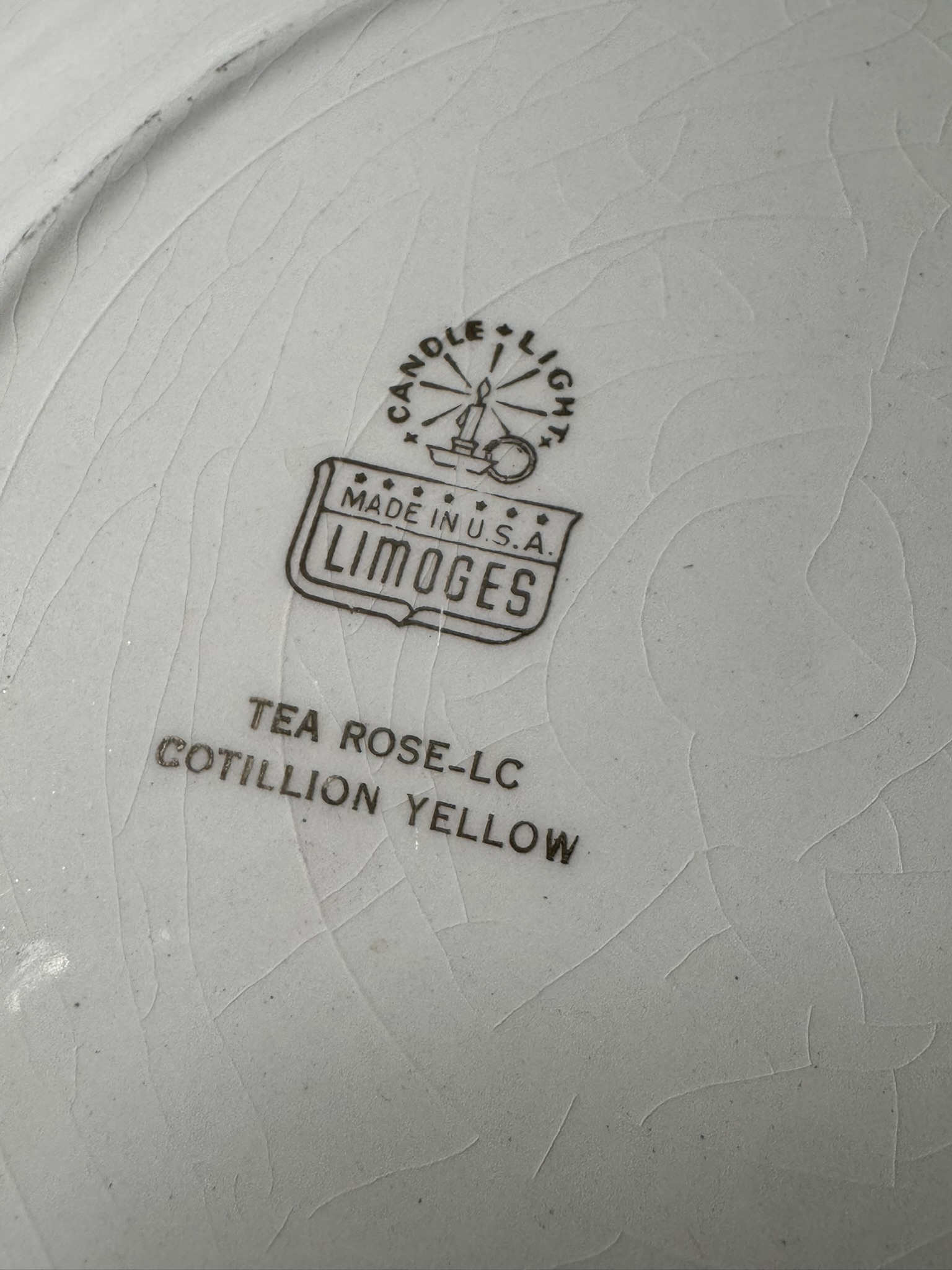 Close-up of the makers mark on a piece of white ceramic with visible crackle texture. The stamp reads “CANDLELIGHT MADE IN U.S.A. LIMOGES,” and below that, “TEA ROSE–LC COTILLION YELLOW” in capital letters.