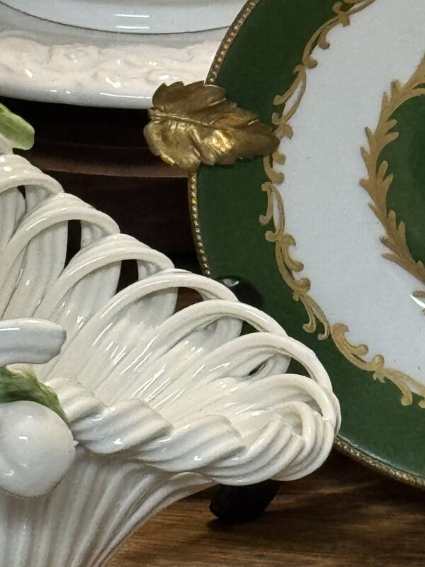Close-up of ornate ceramic items: on the left, a white basket with woven texture and green leaf accents; on the right, a decorative plate with a green border, gold leaf, and scroll details, partly visible, resting on a wooden surface.
