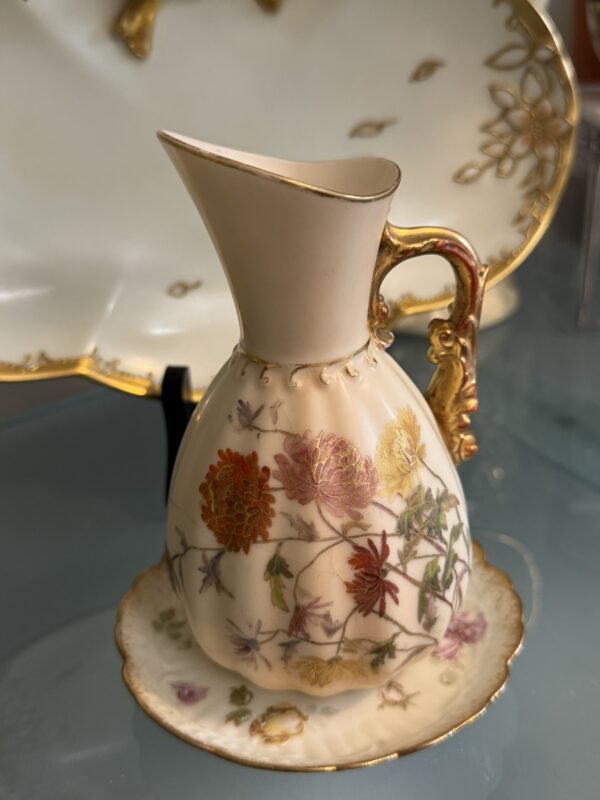 A delicate porcelain pitcher with a gold-trimmed handle and spout sits on a matching saucer. The pitcher is adorned with hand-painted colorful flowers and leaves. A white porcelain dish with gold accents is blurred in the background.