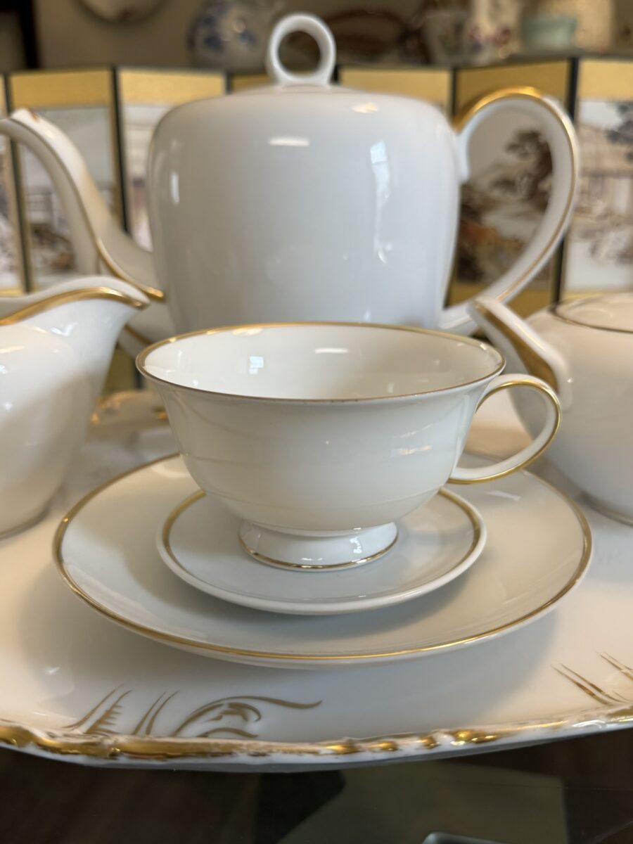 A delicate white porcelain teacup with a matching saucer and gold trim sits on a tray. Behind it are a teapot, creamer, and sugar bowl, all in the same white and gold design. The background has a decorative gold and illustrated screen.
