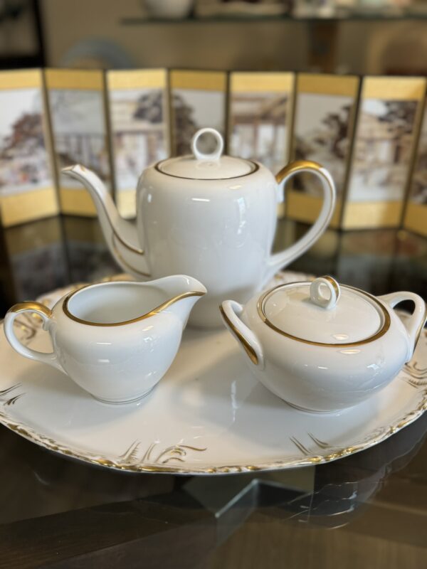 A white porcelain tea set with gold trim sits on a decorative platter. The set includes a teapot, a creamer, and a sugar bowl with a lid. In the background, there is a folding screen with Japanese-style artwork and gold accents.
