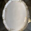 An ornate, oval white porcelain platter with an embossed, scalloped rim and gold decorative accents, sits on a glass tabletop. The platter appears vintage, with intricate gold detailing along the edge and a glossy finish.