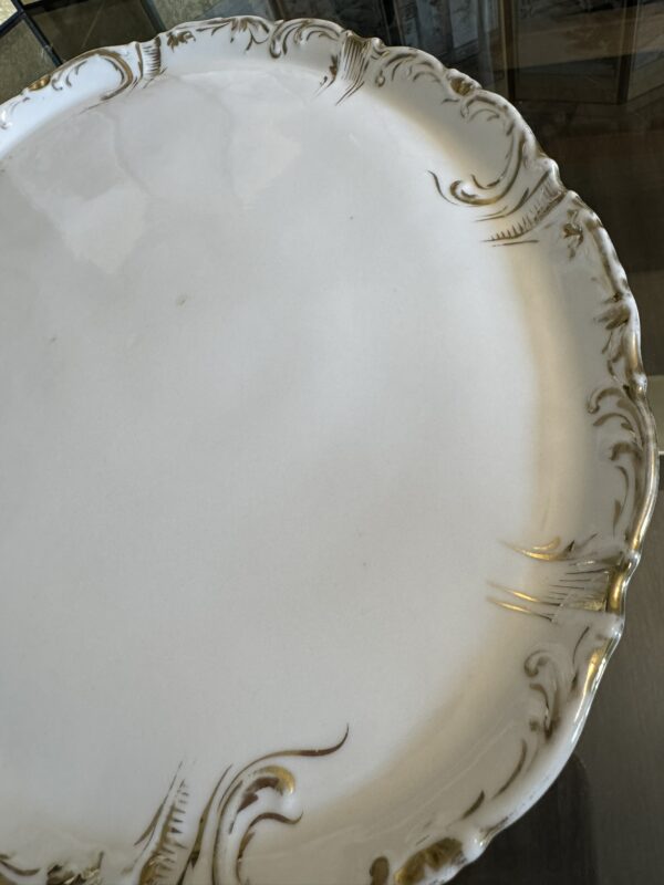 A close-up of an ornate white ceramic plate with scalloped edges and gold decorative accents. The surface is glossy and mostly plain, with raised, swirling patterns along the rim. The plate sits on a glass table.