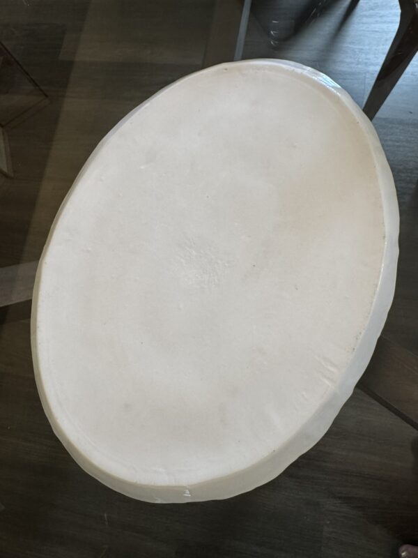 An off-white, oval-shaped tortilla sits atop a glass table, lit by natural light. The tortilla has a smooth, slightly uneven surface and appears thick, with rounded edges. The wooden floor and parts of chairs are visible in the background.