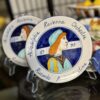 A decorative ceramic plate features a painted profile of a woman in blue and orange attire. The plate reads Philadelphia Ravenna Orchestra, 1992, and Riccardo Muti Cristina. Another similar plate is partially visible behind it.