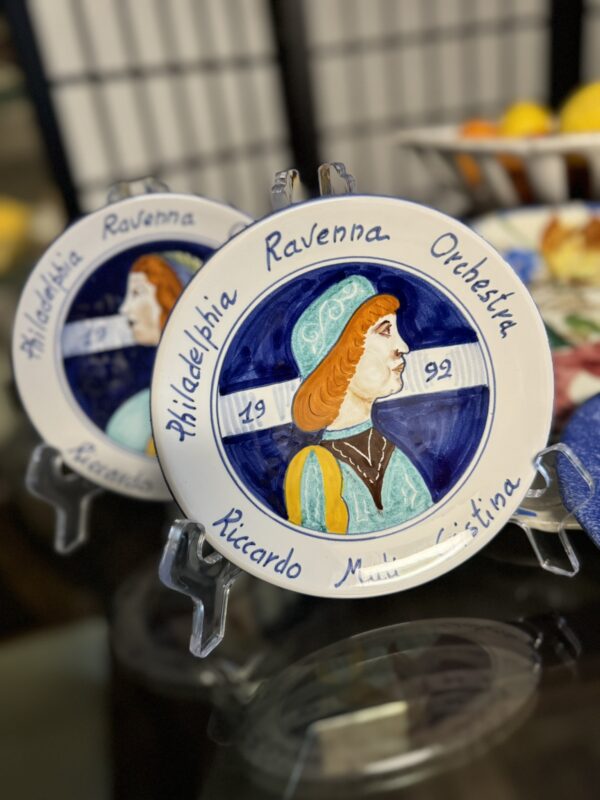 A decorative ceramic plate features a painted profile of a woman in blue and orange attire. The plate reads Philadelphia Ravenna Orchestra, 1992, and Riccardo Muti Cristina. Another similar plate is partially visible behind it.