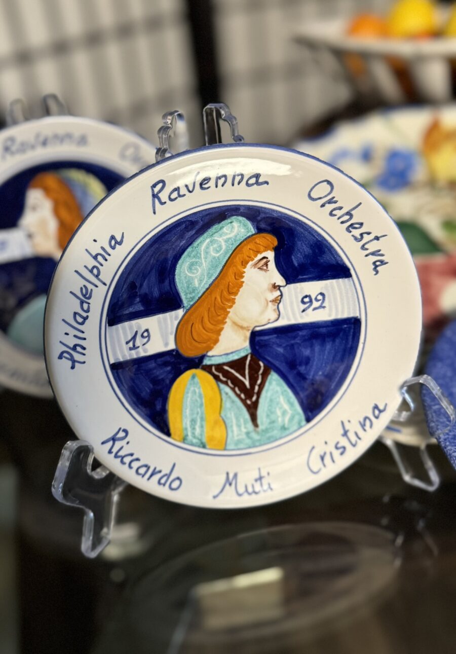 A decorative ceramic plate on a stand features a painted profile of a woman in Renaissance attire. The plate reads “Philadelphia Ravenna Orchestra 1992 Riccardo Muti Cristina” in blue script around the edge. A similar plate is partially visible behind it.