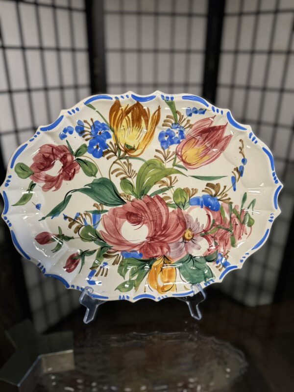 A decorative ceramic platter with scalloped edges outlined in blue, featuring a hand-painted floral design with large red, yellow, and pink flowers, green leaves, and small blue blossoms, displayed upright on a clear stand against a shoji screen background.