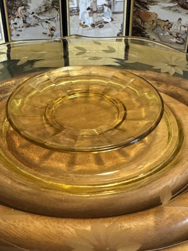A transparent amber glass plate rests on stacked wooden trays, with a decorative screen in the background showing painted scenes of people and animals in traditional attire and settings. The glass plate reflects warm light, highlighting its smooth, circular shape.