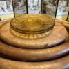 A round amber glass plate sits on stacked circular wooden boards. Behind them is a folding screen featuring traditional Asian-style art with people, trees, and buildings in soft tones. Warm lighting highlights the textures of the wood and glass.