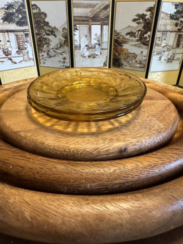 A round amber glass plate sits on stacked circular wooden boards. Behind them is a folding screen featuring traditional Asian-style art with people, trees, and buildings in soft tones. Warm lighting highlights the textures of the wood and glass.