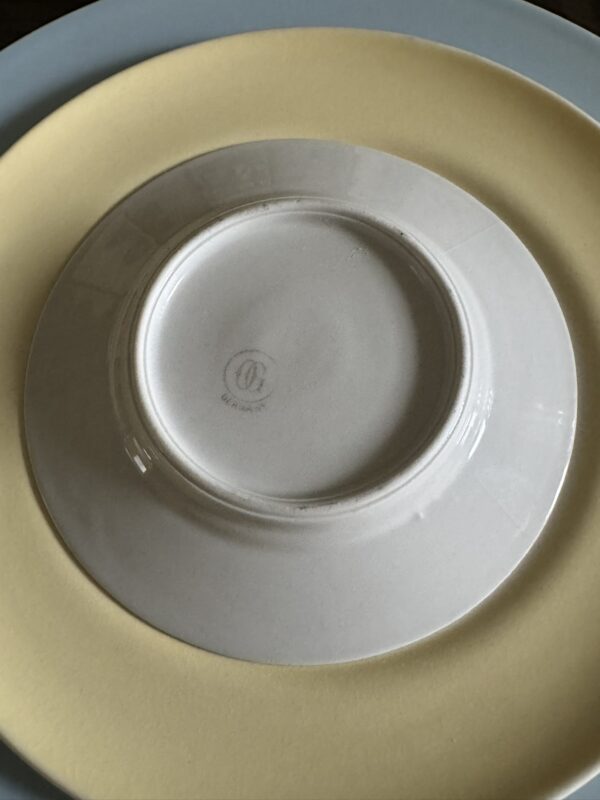 A close-up of three stacked plates: the top plate is white and upside down, showing a faint manufacturer’s mark at its center; the middle plate is pale yellow, and the bottom plate is pale blue, both visible as circular borders beneath the white plate.