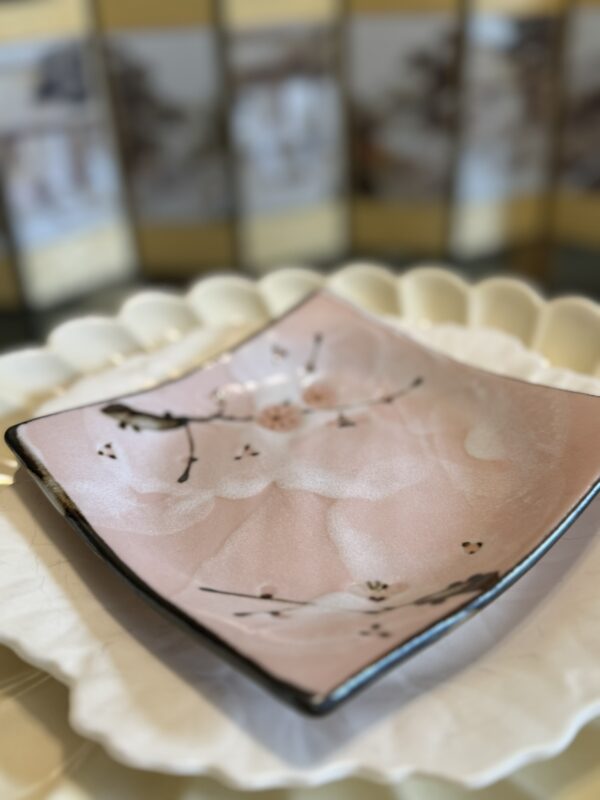 A square, pink ceramic plate with a delicate cherry blossom design sits atop a larger, white, scalloped-edge plate. In the background, there’s a decorative screen with muted images, softly out of focus. The overall scene feels elegant and serene.