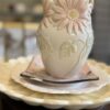 A cream-colored ceramic vase with raised pink daisy flowers and green leaves sits on stacked plates: a white scalloped plate and a square plate with a pink and black design, set on a round wooden surface with a softly blurred background.