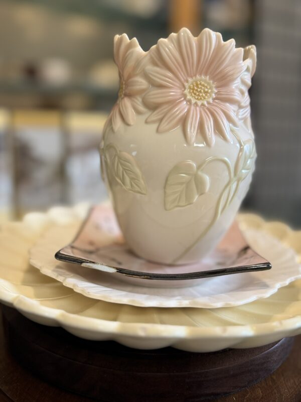 A cream-colored ceramic vase with raised pink daisy flowers and green leaves sits on stacked plates: a white scalloped plate and a square plate with a pink and black design, set on a round wooden surface with a softly blurred background.